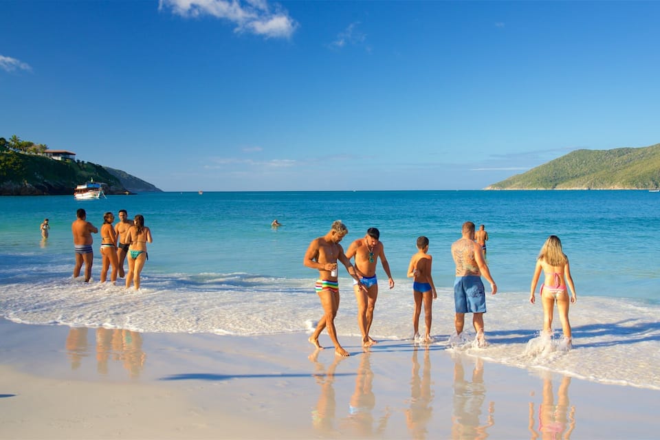 A beach with people walking along the shore, a boat in the water, and hills in the background.