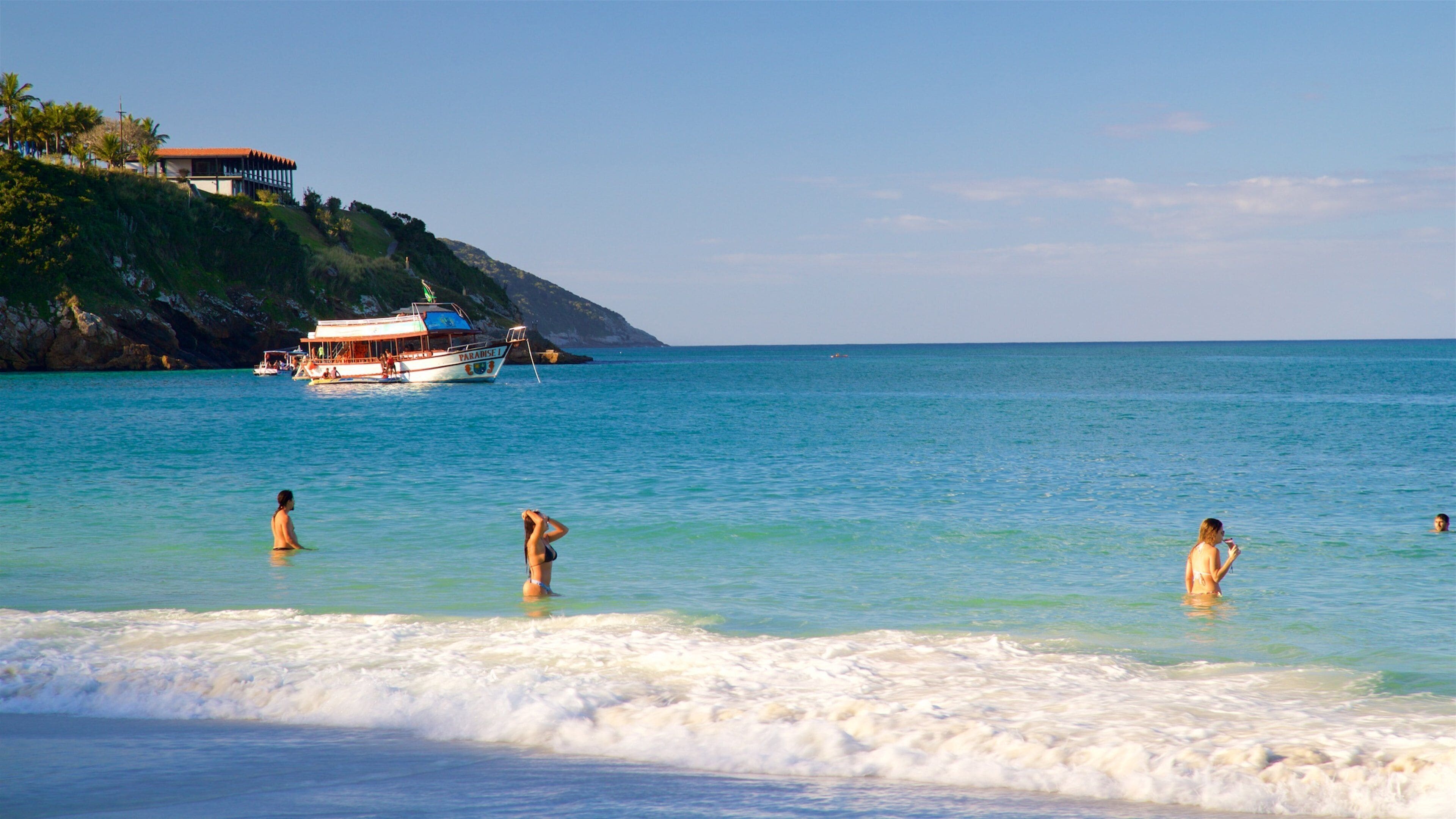 Une plage à l’eau turquoise et limpide, un bateau baptisé « Paradise I » ancré près du rivage, et des gens qui se baignent et prennent des bains de soleil.