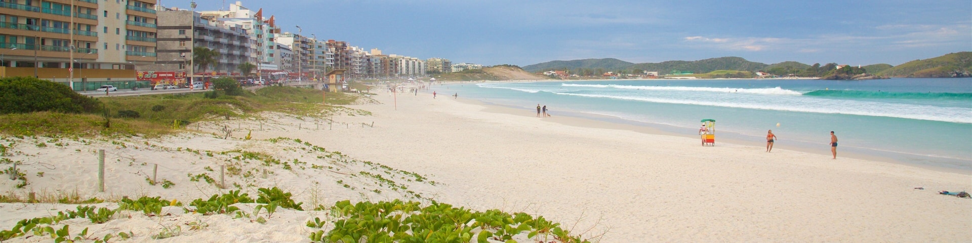 Una playa arenosa con olas, un puesto de salvavidas y edificios al fondo.