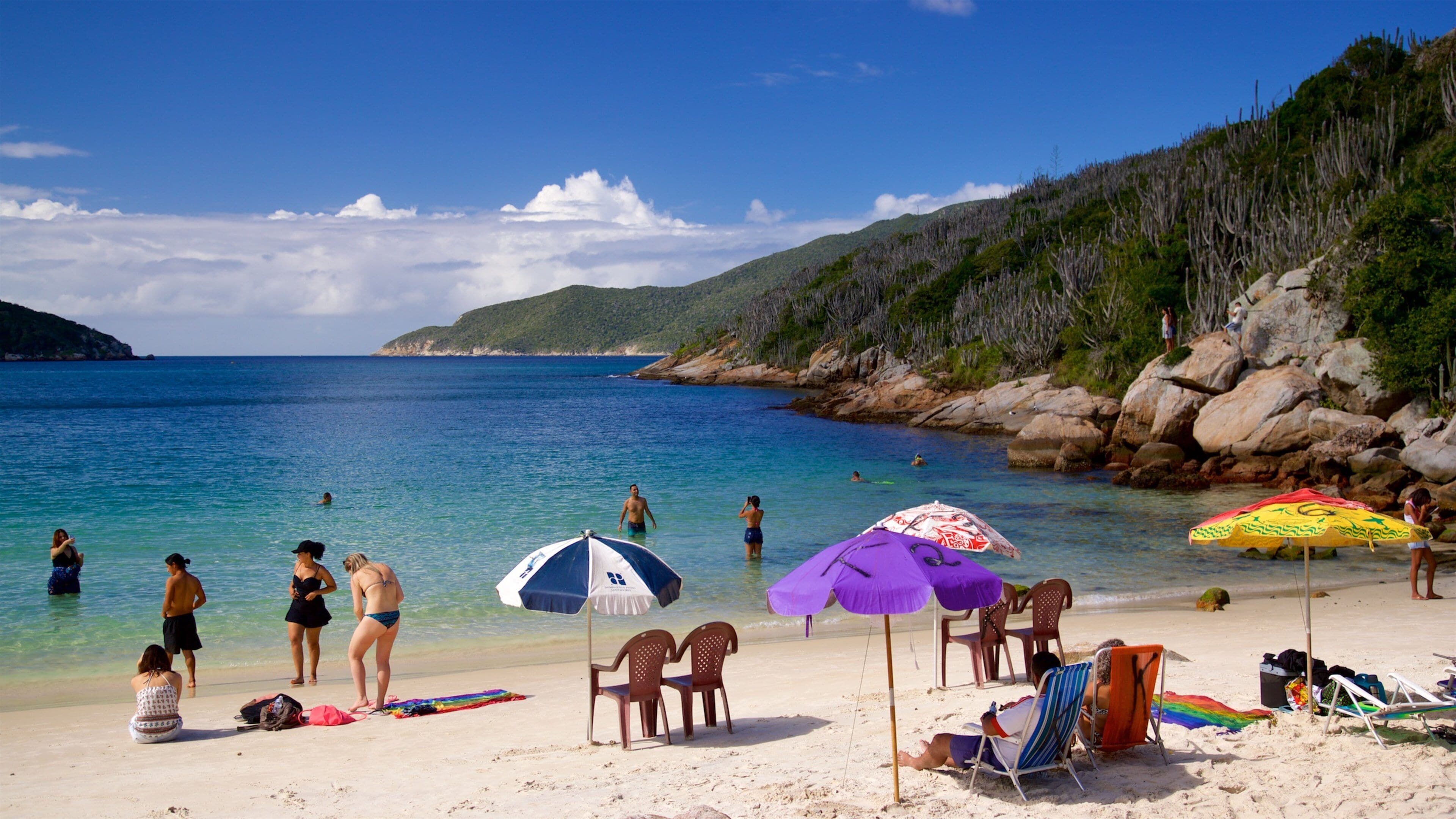 Une plage avec une eau turquoise et limpide, des gens qui prennent un bain de soleil et des parasols.