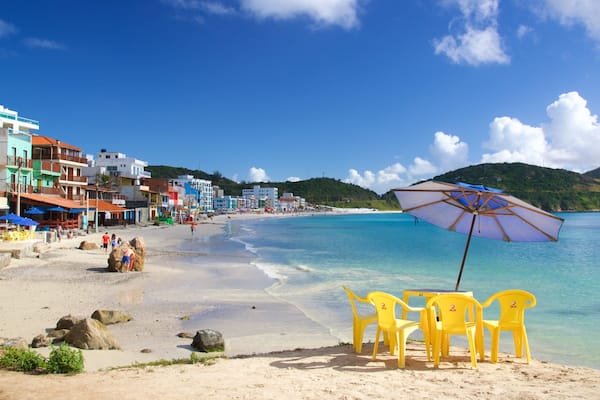 Uma praia com águas turquesa cristalinas, edifícios coloridos e uma orla arenosa com cadeiras amarelas e um guarda-sol.