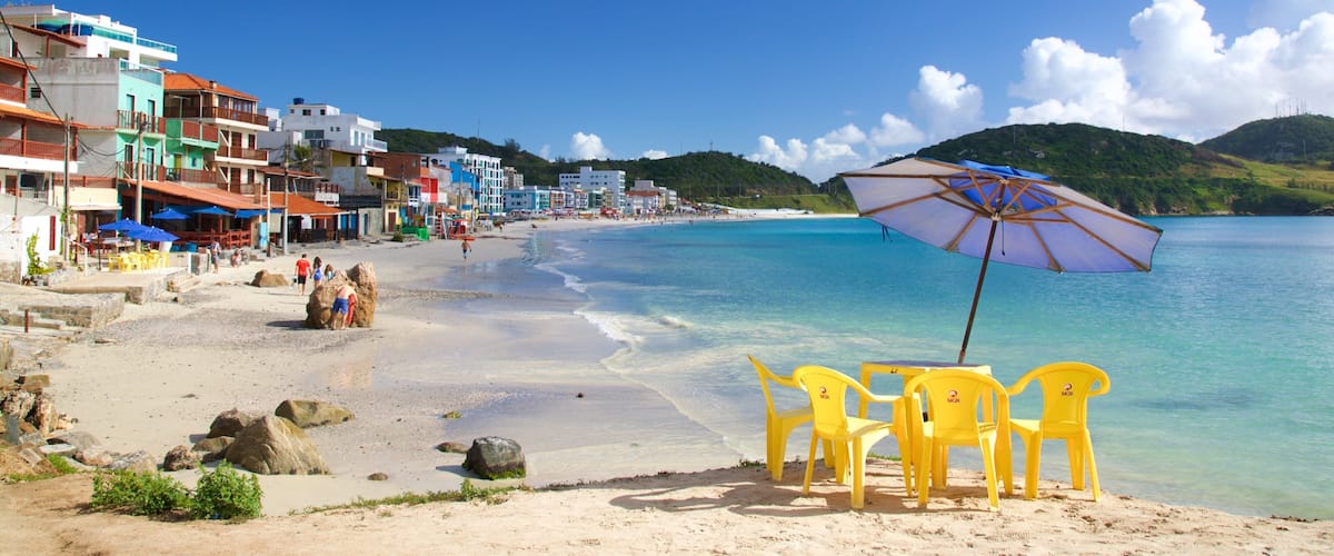 A beach with clear turquoise water, colorful buildings, and a sandy shore with yellow chairs and an umbrella.