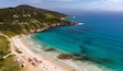 A coastal landscape with a beach, clear turquoise waters, and a hilly backdrop.