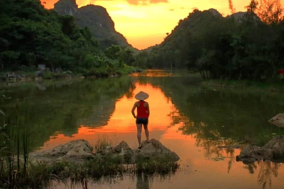 A person in a red top and hat standing by a calm river at sunset.