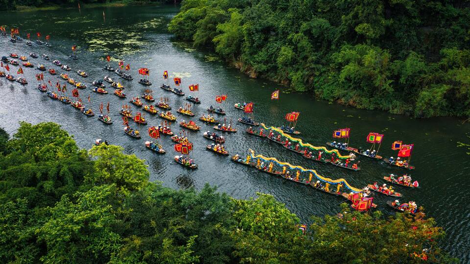 A river with numerous boats adorned with colorful decorations and flags, surrounded by dense greenery.