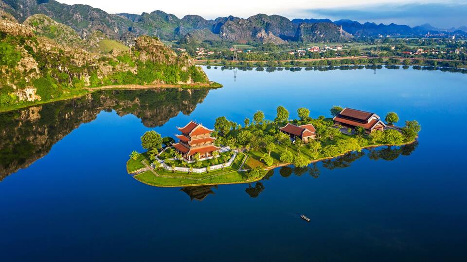 An aerial view of a traditional temple complex on a small island surrounded by a lake, with mountains in the background.