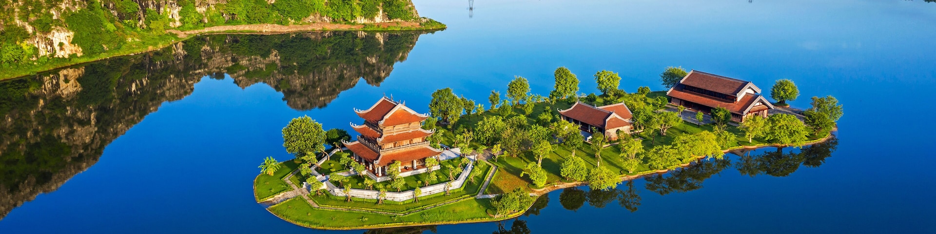 An aerial view of a traditional temple complex on a small island surrounded by a lake, with mountains in the background.