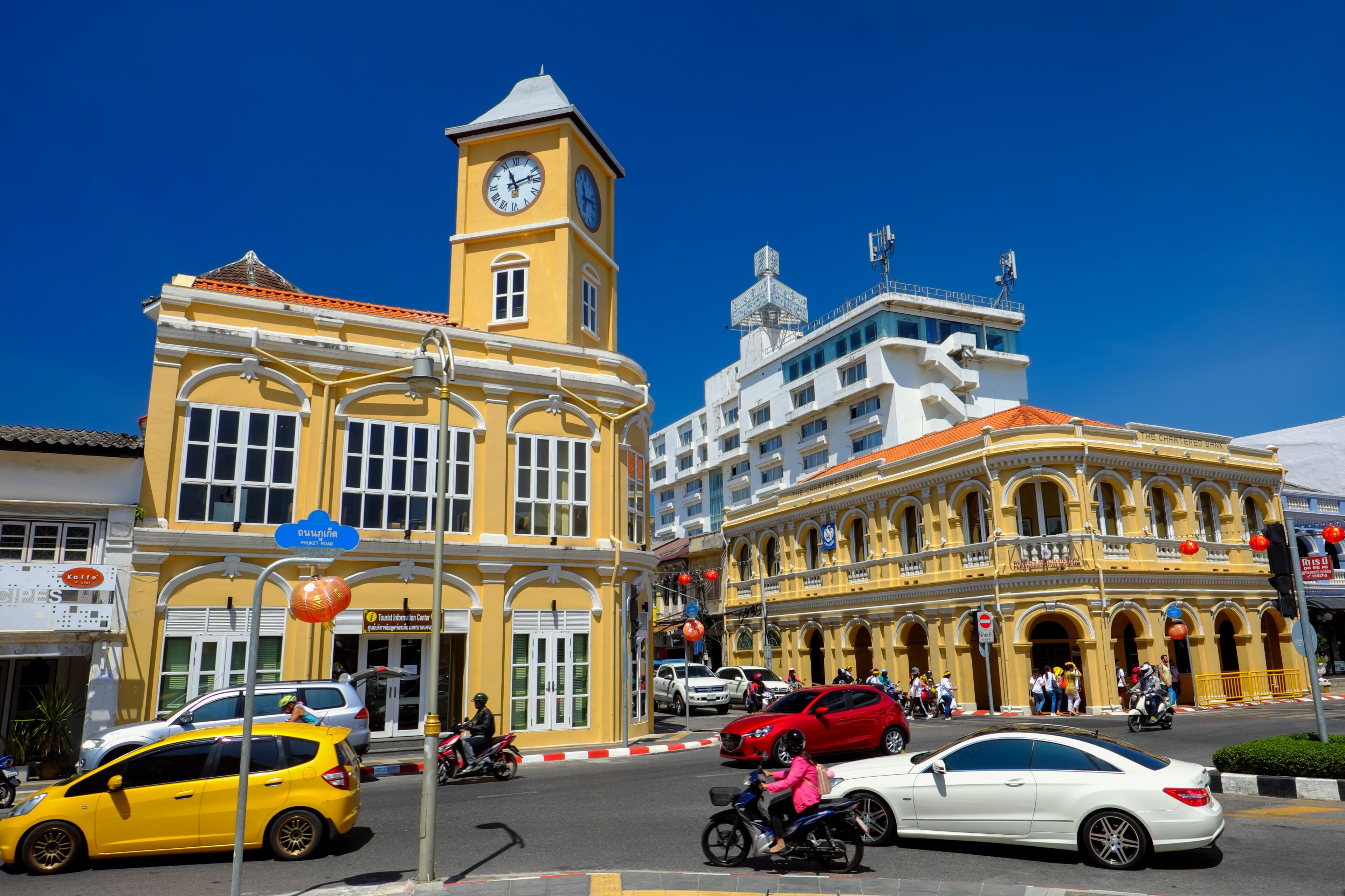 A yellow building with a clock tower, a white building with balconies, and a street with cars and motorbikes.