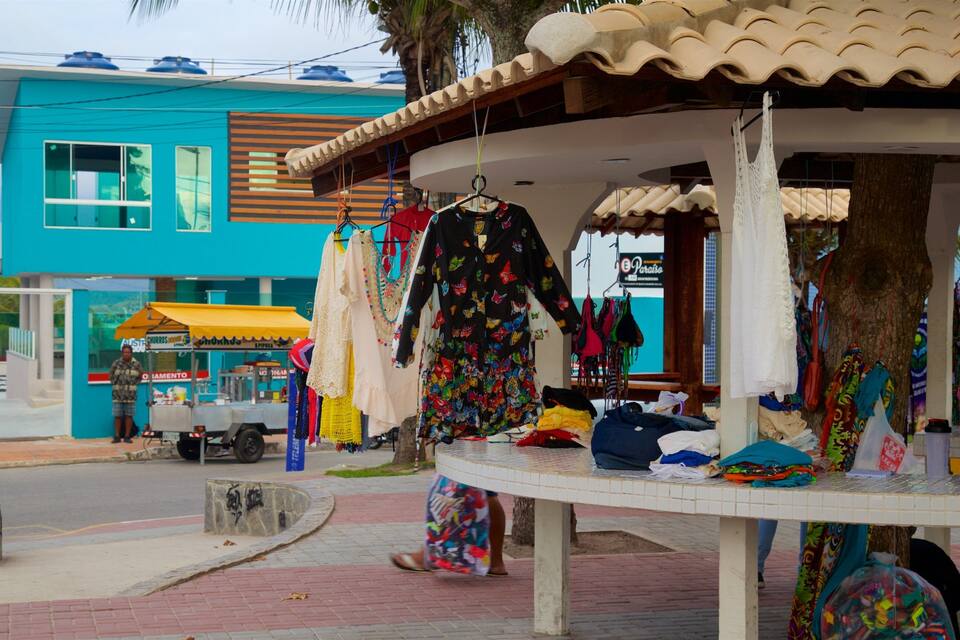 Uma banca de roupas colorida com diversas peças penduradas, com um edifício azul e um food truck estacionado ao fundo.
