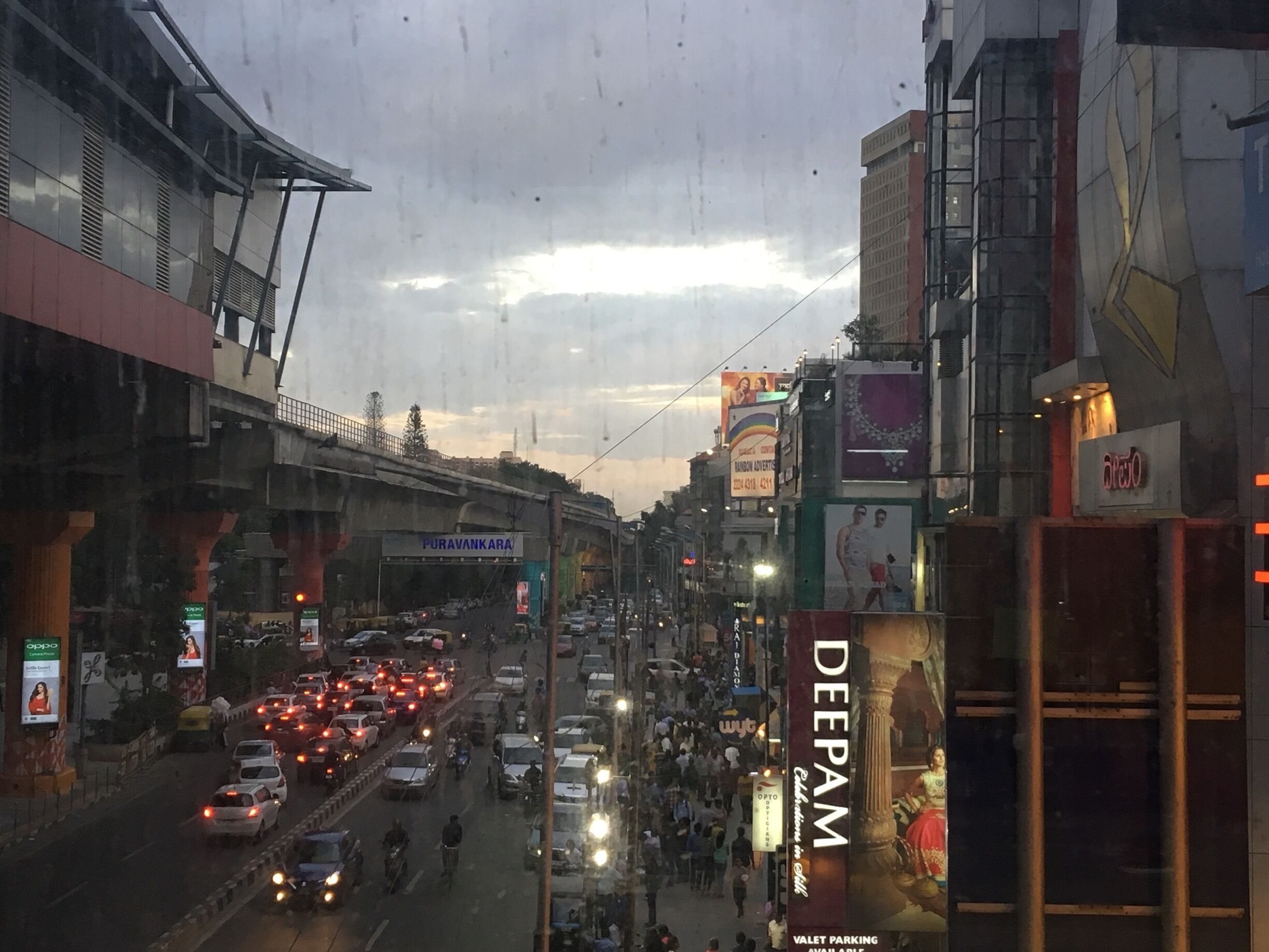 Uma rua urbana movimentada com tráfego intenso, outdoors e uma ponte sobre ela.