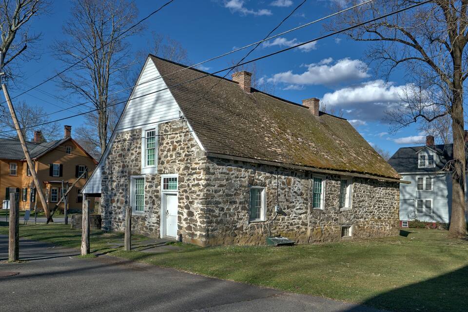 A stone house with a steep gabled roof, surrounded by trees and a clear blue sky.