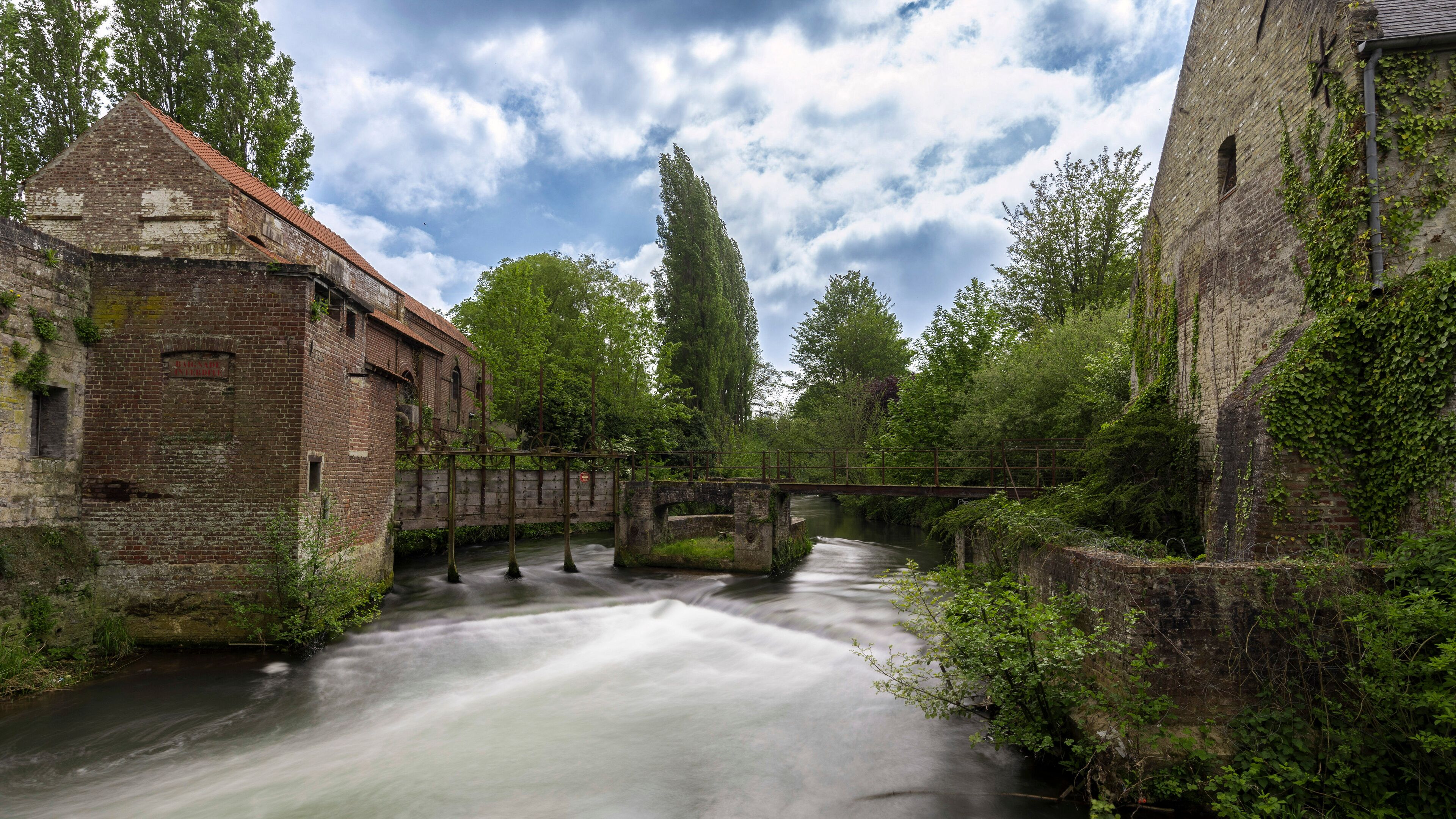 A river flowing through an old brick building with a bridge and lush greenery.