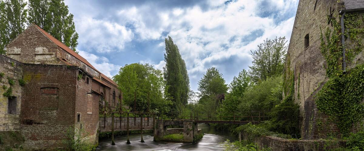 A river flowing through an old brick building with a bridge and lush greenery.