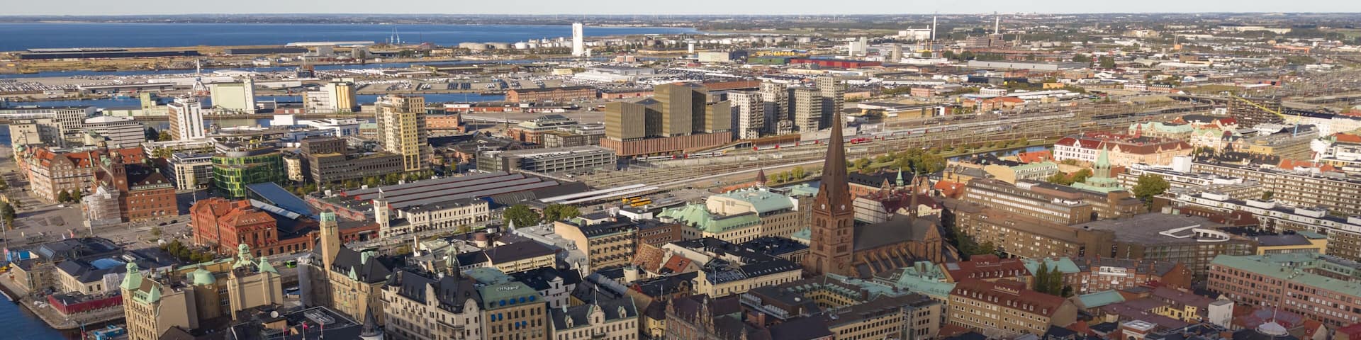 A panoramic view of a historic city with dense architecture, a prominent church spire, and a waterfront in the distance.