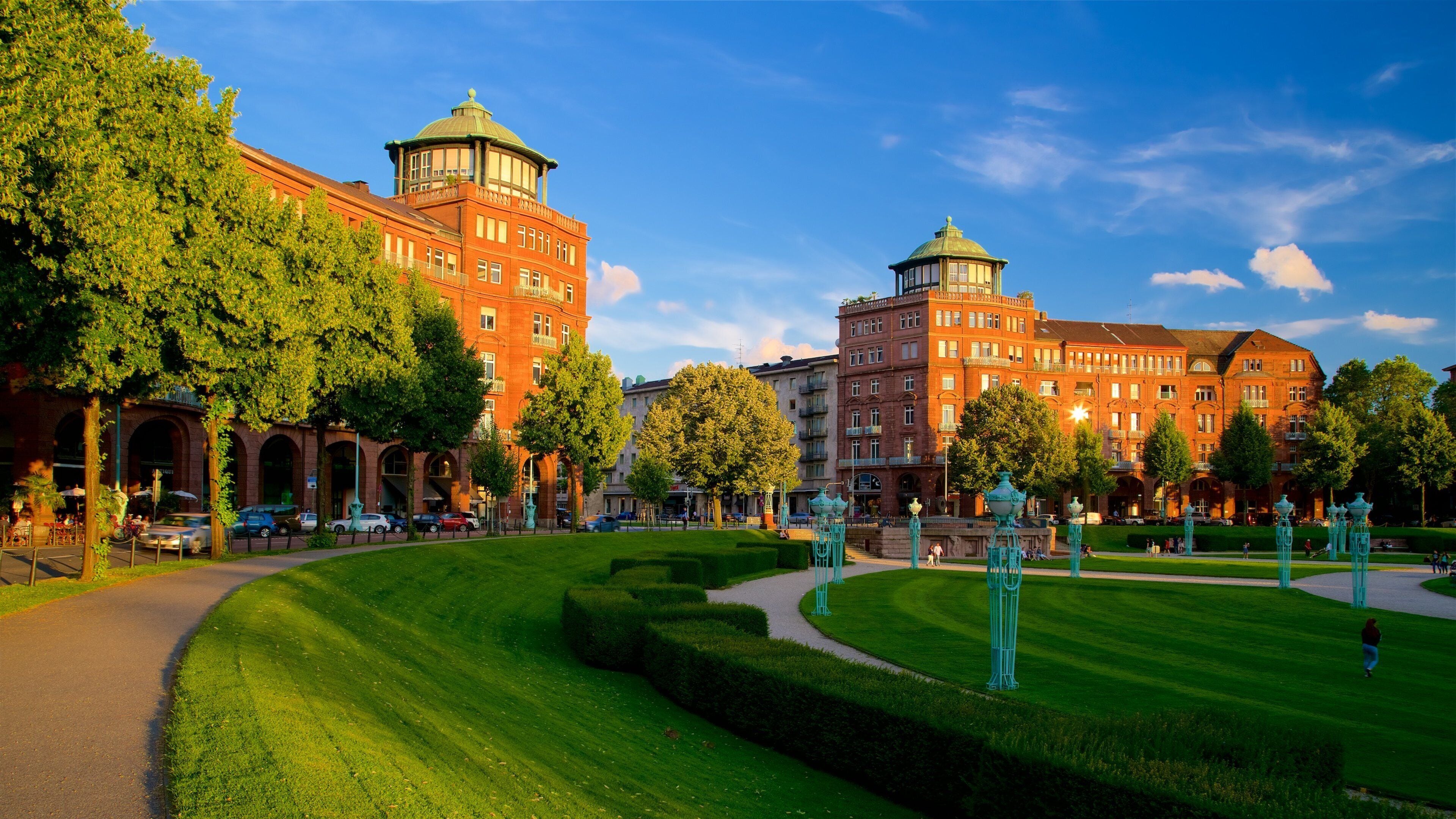 A well-manicured park with a red brick building featuring two domed towers, surrounded by lush green trees and a clear blue sky.