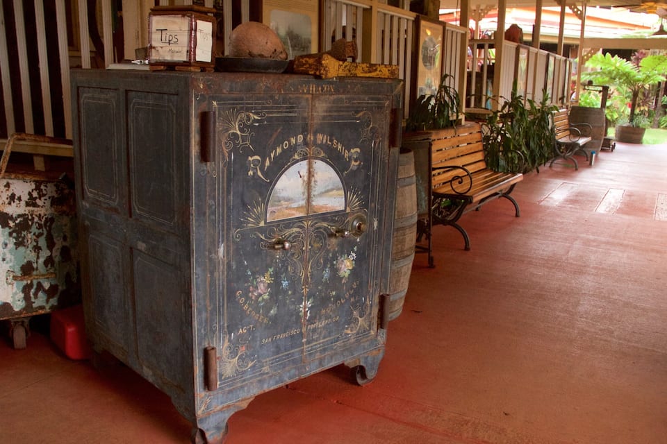 An antique blue cabinet with ornate designs and a glass window, placed outdoors under a shelter with a bench and potted plants nearby.