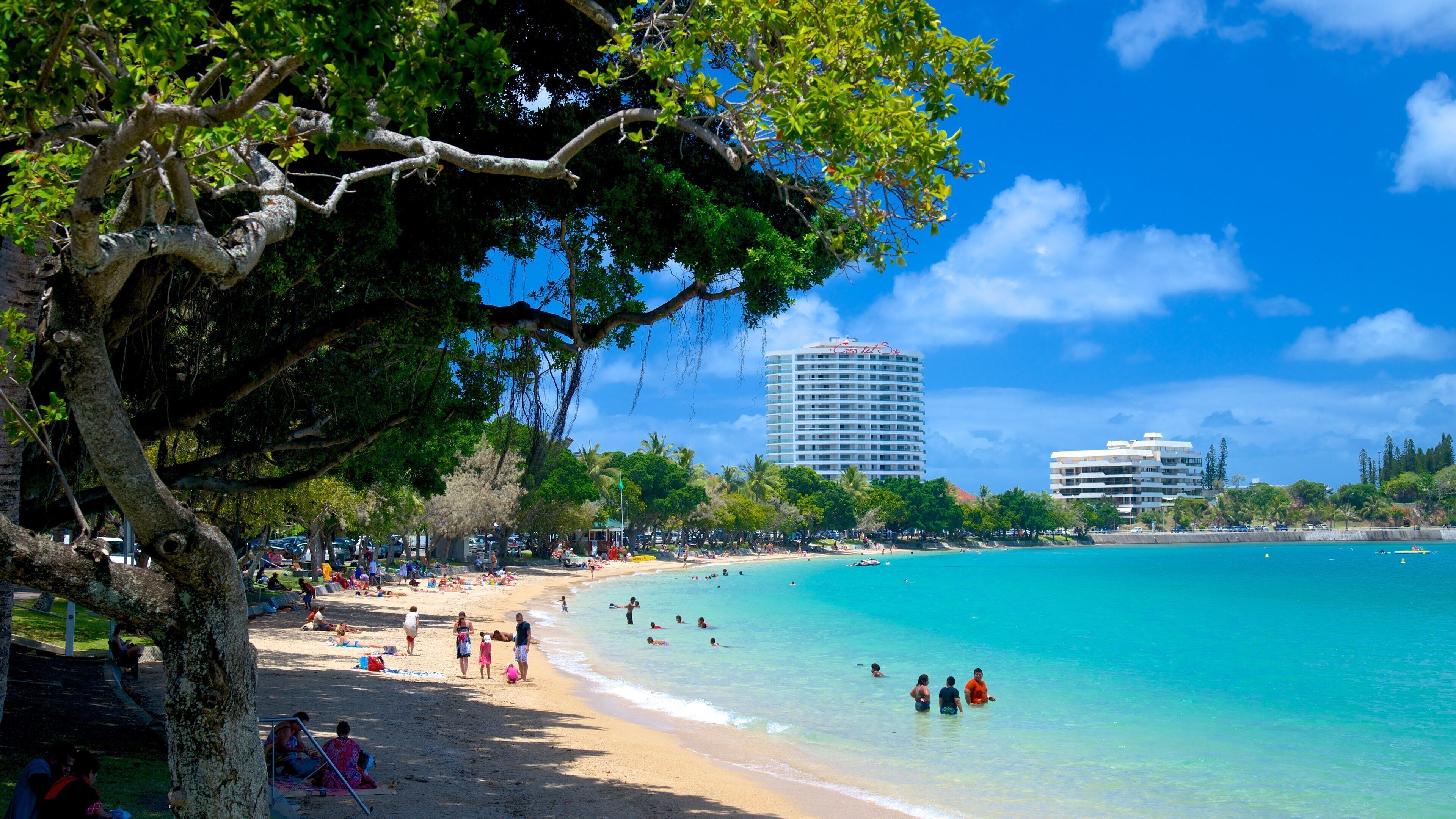 A beach with clear turquoise water, people sunbathing, and tall buildings in the background.