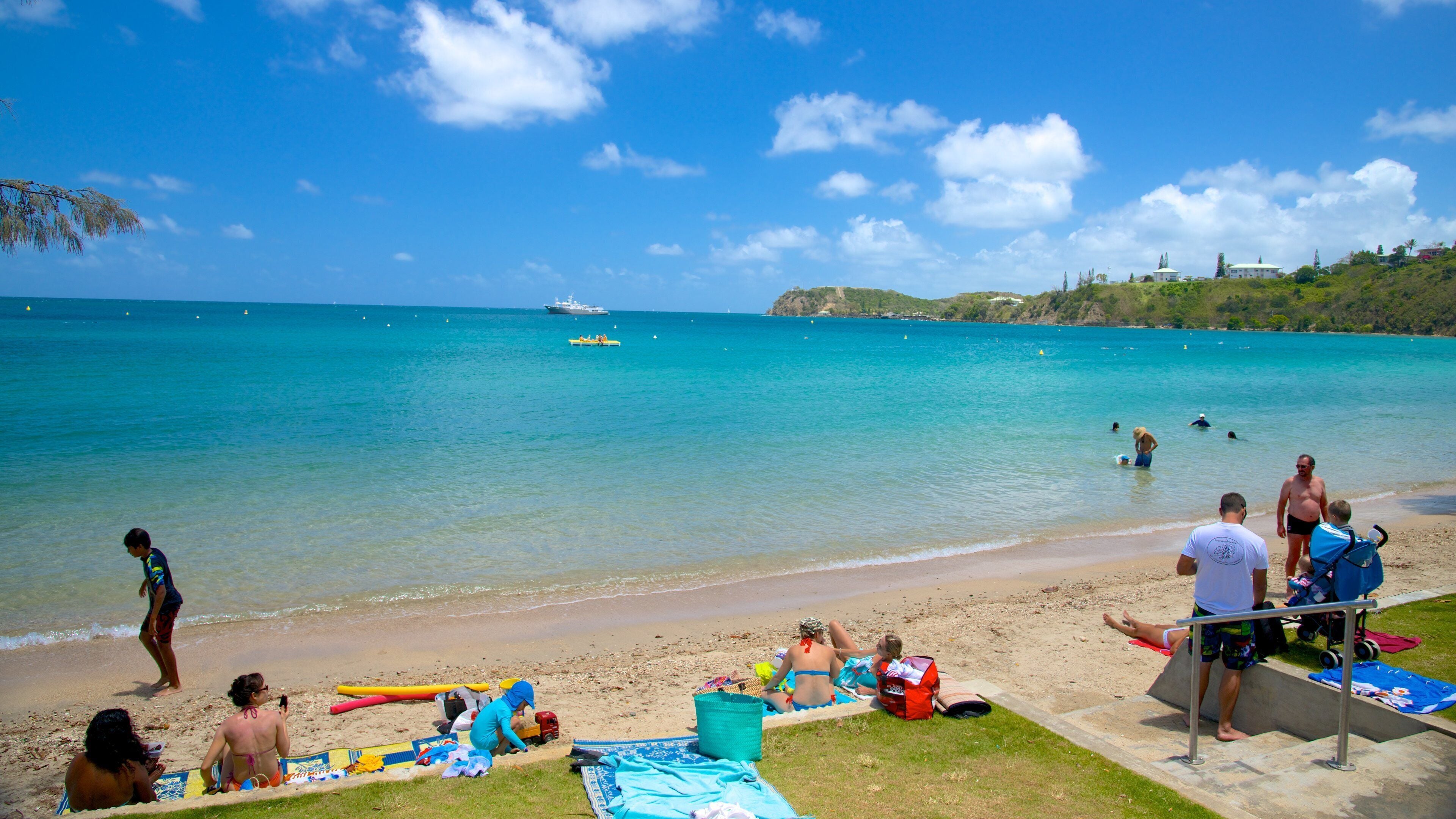 A beach with clear turquoise water, people sunbathing, and a ship in the distance.