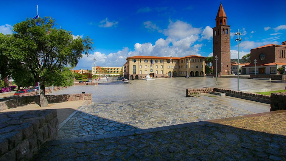 A plaza with a prominent clock tower, a large building with a red roof, and a tree in the foreground.