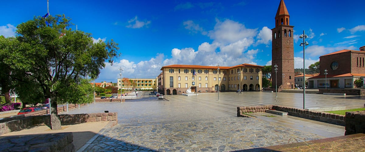 A plaza with a prominent clock tower, a large building with a red roof, and a tree in the foreground.