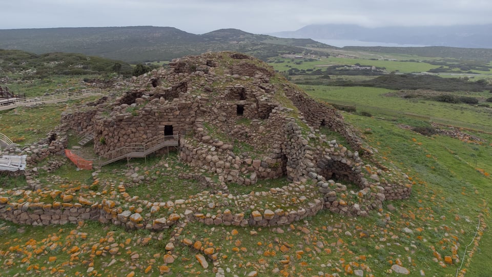 A stone ruin with a staircase and a small opening, surrounded by green fields and mountains in the background.