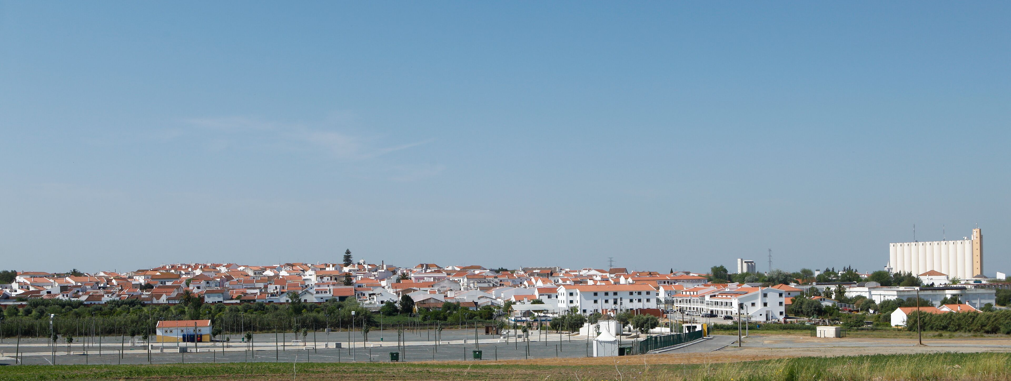A coastal town with red-roofed houses, a large grain silo, and a clear blue sky.