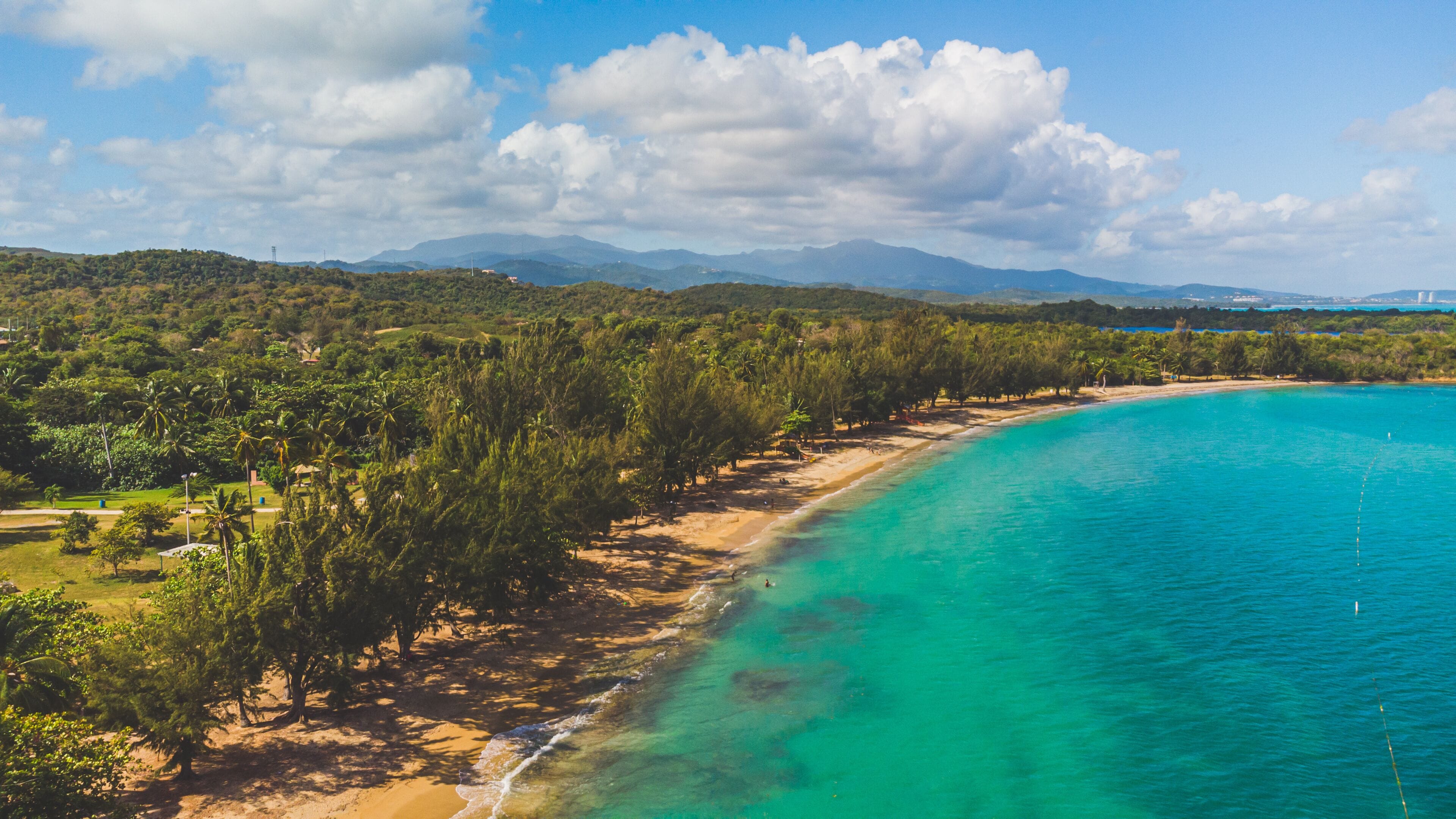 A coastal landscape with a sandy beach, turquoise waters, dense greenery, and distant mountains.