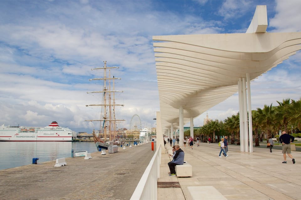 A cruise ship docked at a port with a large sailboat nearby, a Ferris wheel in the background, and a modern white structure with a curved roof.