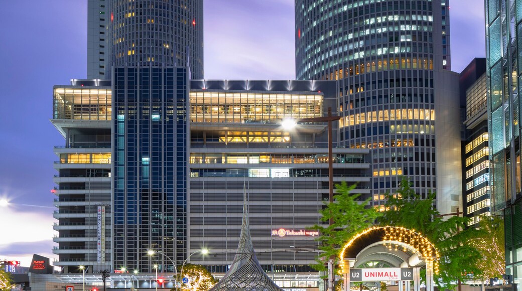 A cityscape at dusk with two prominent skyscrapers, a modern building with a spire, and a busy street with light trails from moving vehicles.