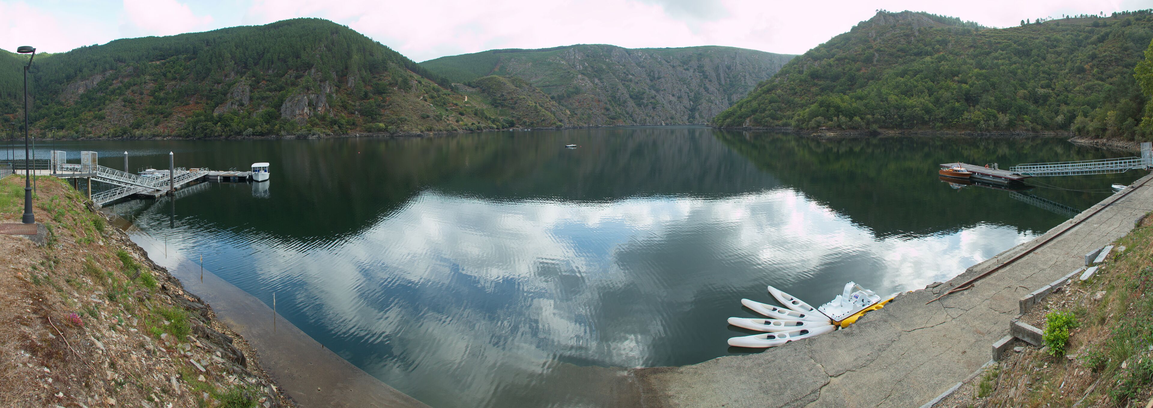 A calm lake with boats docked at a pier, surrounded by lush green hills.