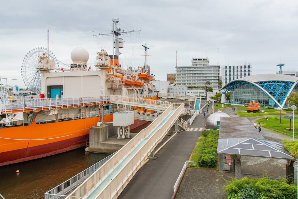 A large orange ship docked at a port with a Ferris wheel and modern buildings in the background.