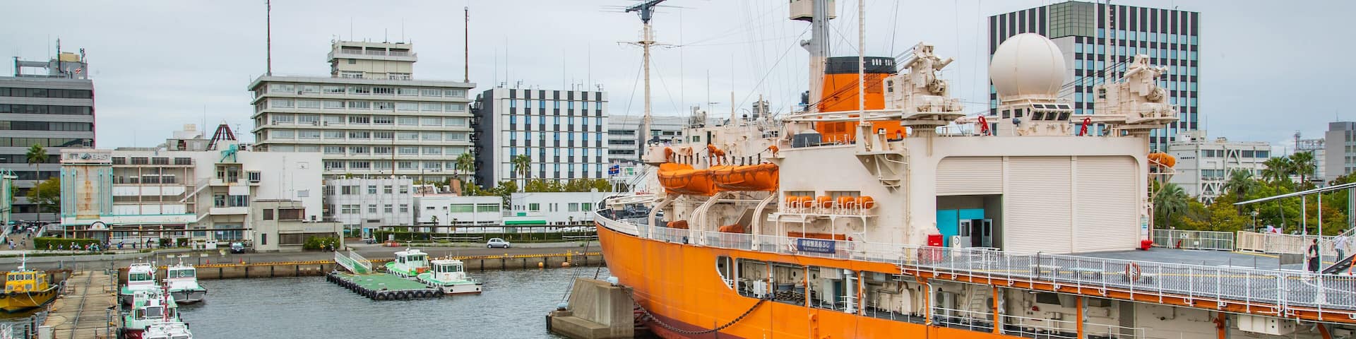 A large orange ship docked at a port with a city skyline in the background.