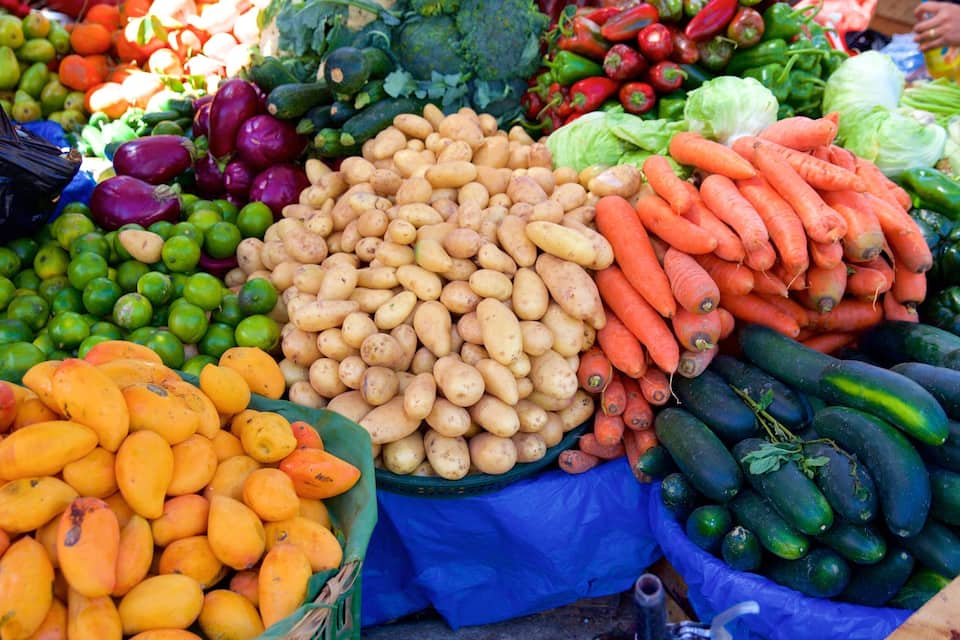 Variedad de verduras frescas, incluyendo papas, zanahorias y pepinos, exhibidas en un mercado.