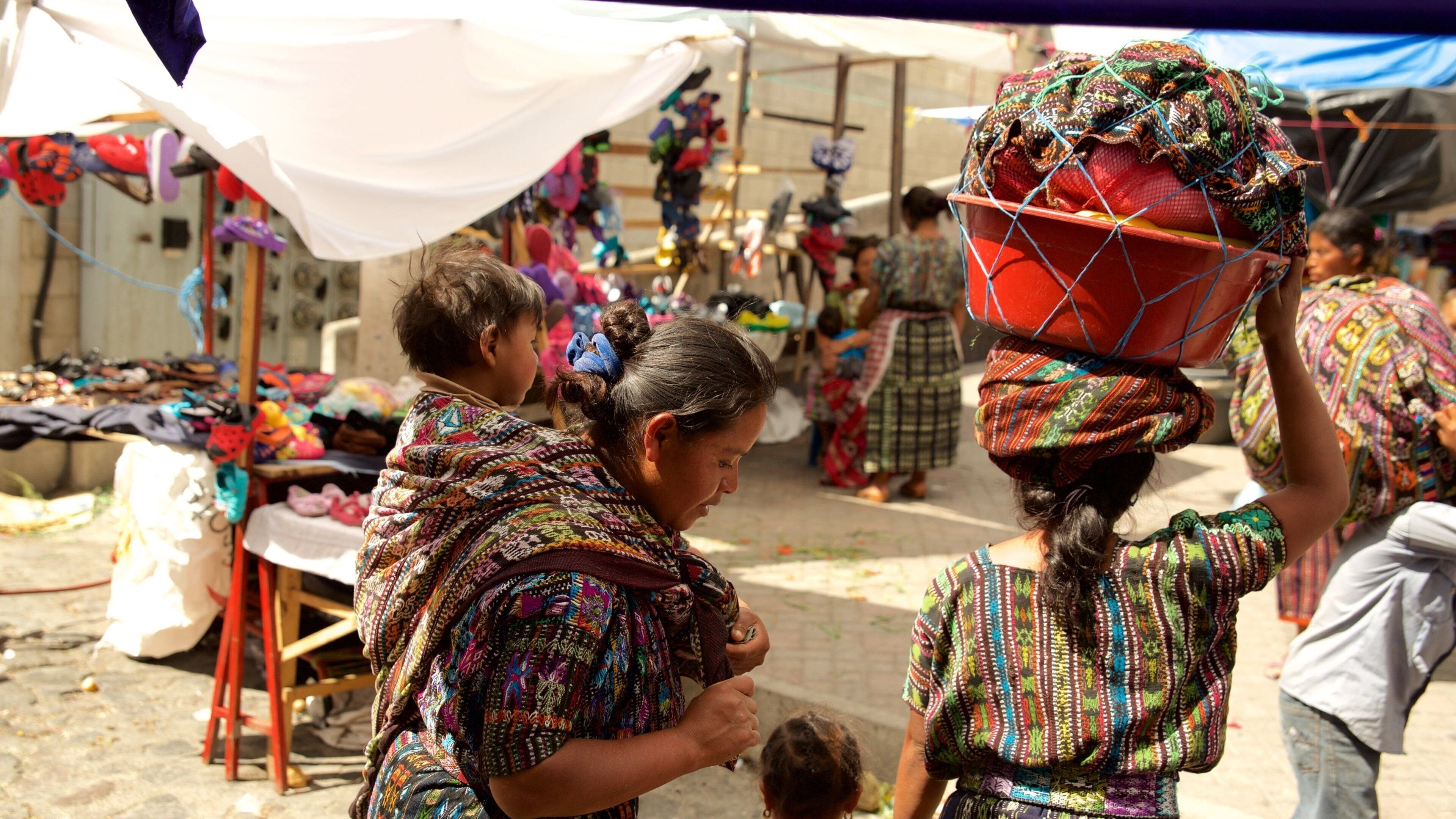 Une scène de marché où l’on voit des personnes portant des marchandises sur leur tête, des étoffes aux couleurs vives et divers articles mis en vente.