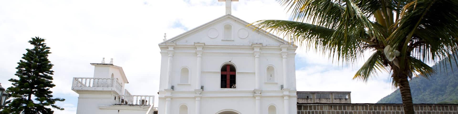Une église blanche surmontée d’une croix, entourée de palmiers et avec une montagne en arrière-plan.