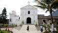A white church with a cross on top, surrounded by palm trees and a mountain in the background.