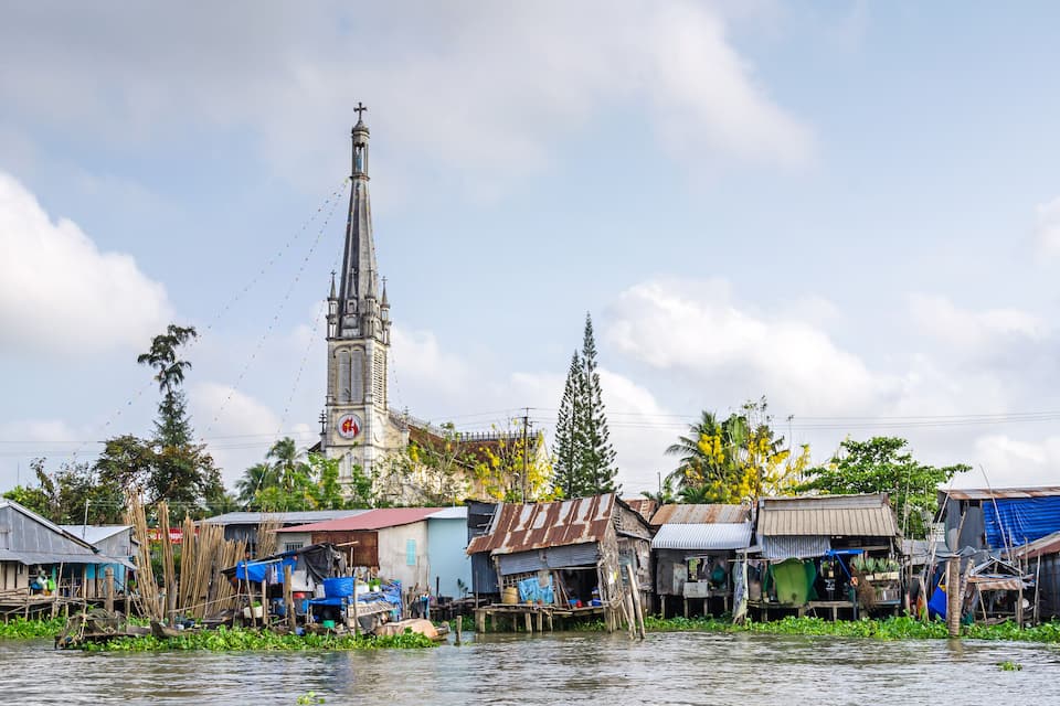 A riverside settlement with makeshift houses, a church with a tall steeple, and lush greenery.