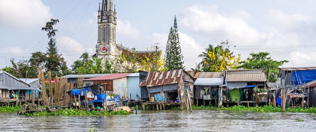 A riverside settlement with makeshift houses, a church with a tall steeple, and lush greenery.