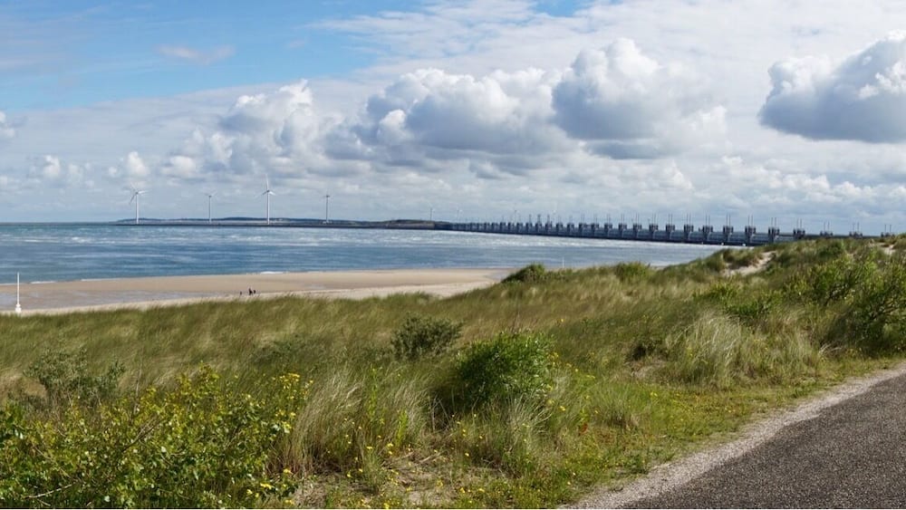 Une route côtière bordée d’éoliennes, une plage et un pont qui se dessine à l’horizon.