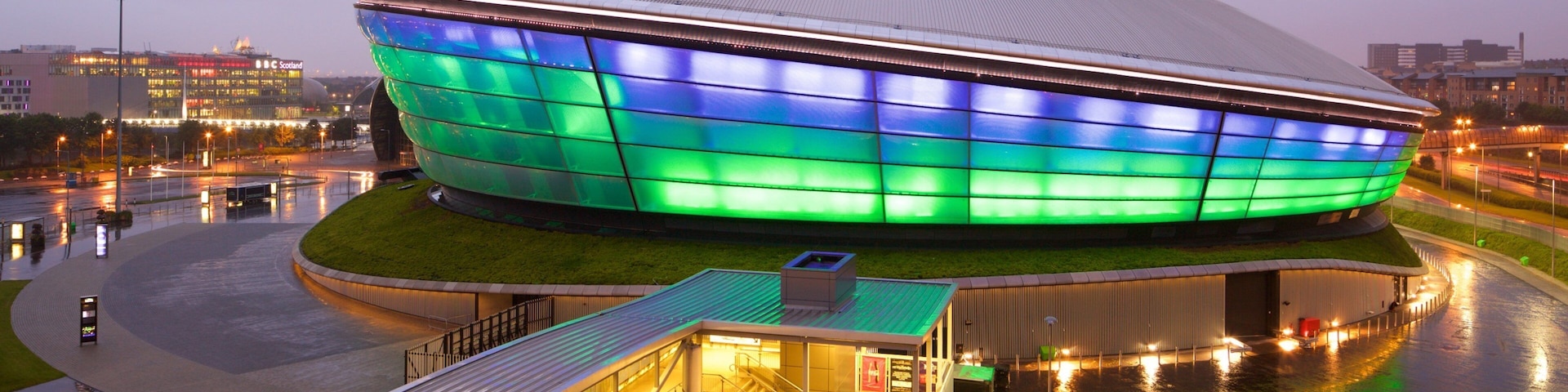 Estádio moderno com um grande teto curvo e iluminação vibrante em tons de verde e azul.