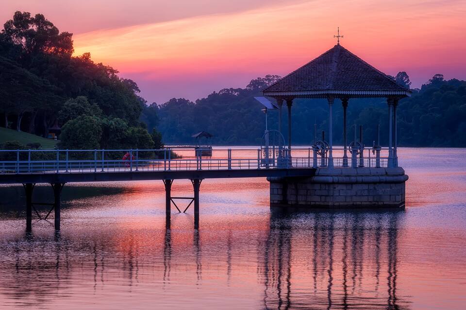 A gazebo on a pier over calm water, surrounded by trees and a sunset sky.