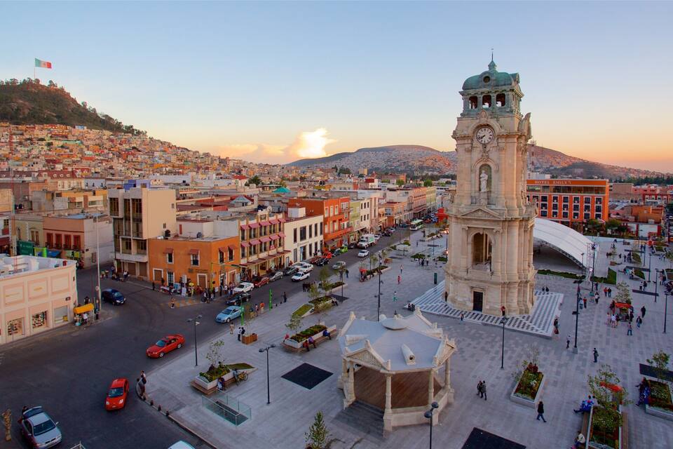 Un paisaje urbano con una destacada torre del reloj, una plaza animada y una colina con edificios.