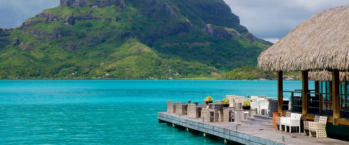 A turquoise sea with a wooden pier, thatched-roof hut, and a mountain in the background.