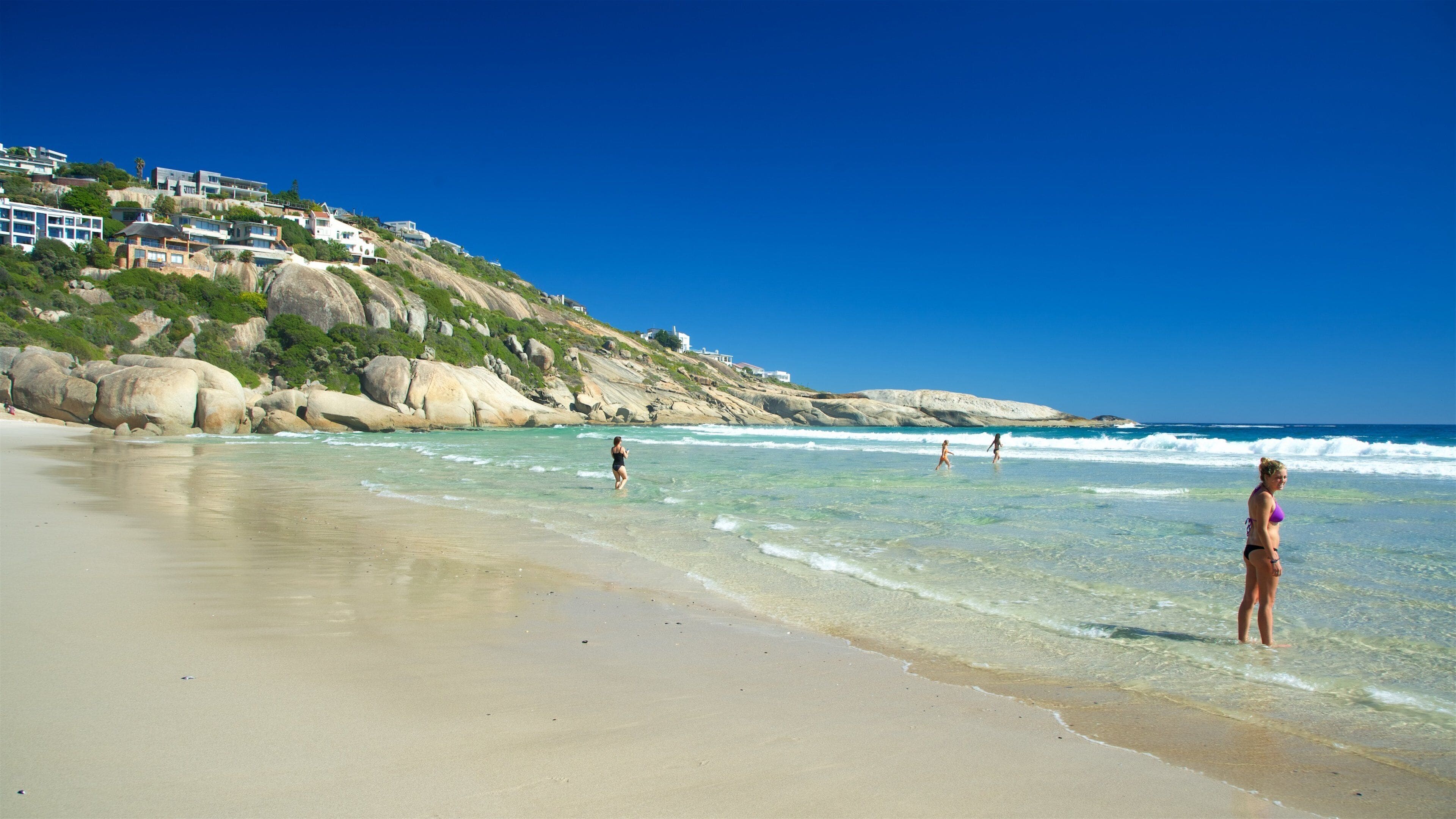 Uma praia com águas turquesa cristalinas, uma faixa de areia e casas numa encosta ao fundo.