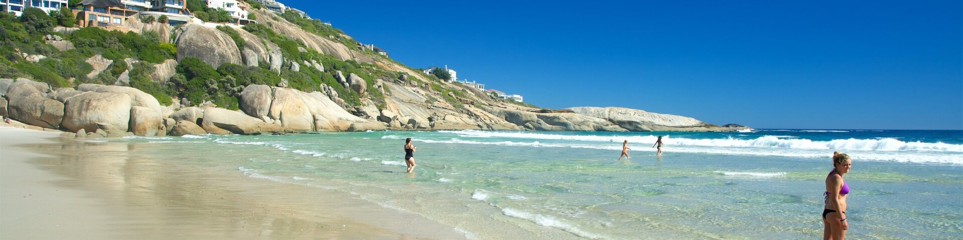 Uma praia com águas turquesa cristalinas, uma faixa de areia e casas numa encosta ao fundo.
