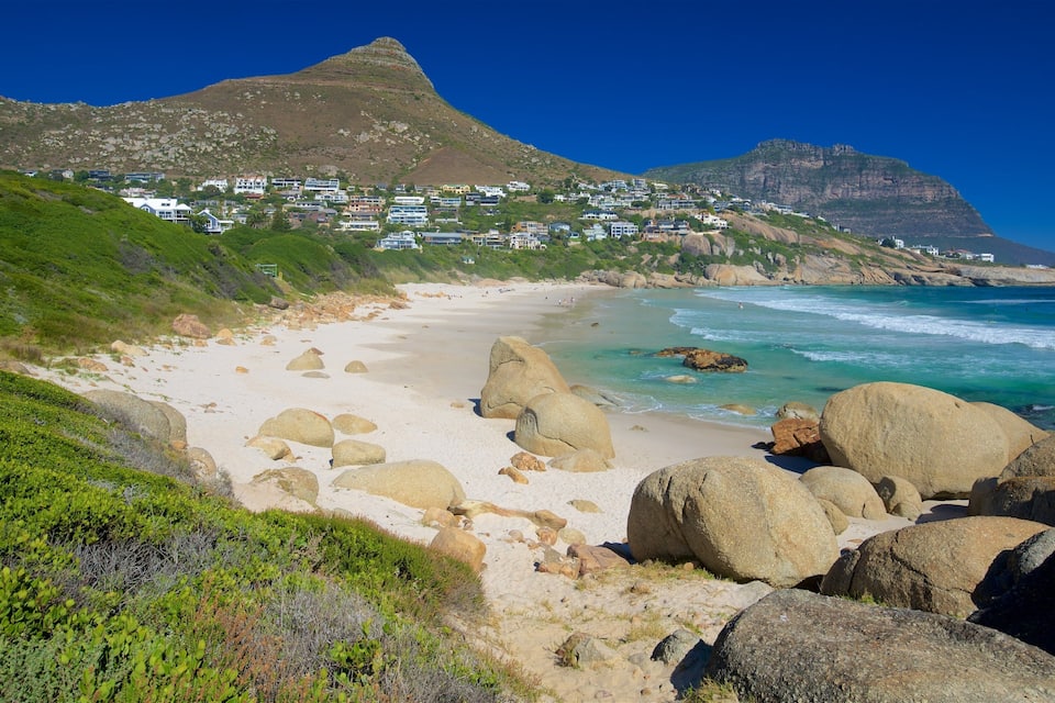A coastal landscape with a sandy beach, large boulders, and a town on a hillside.