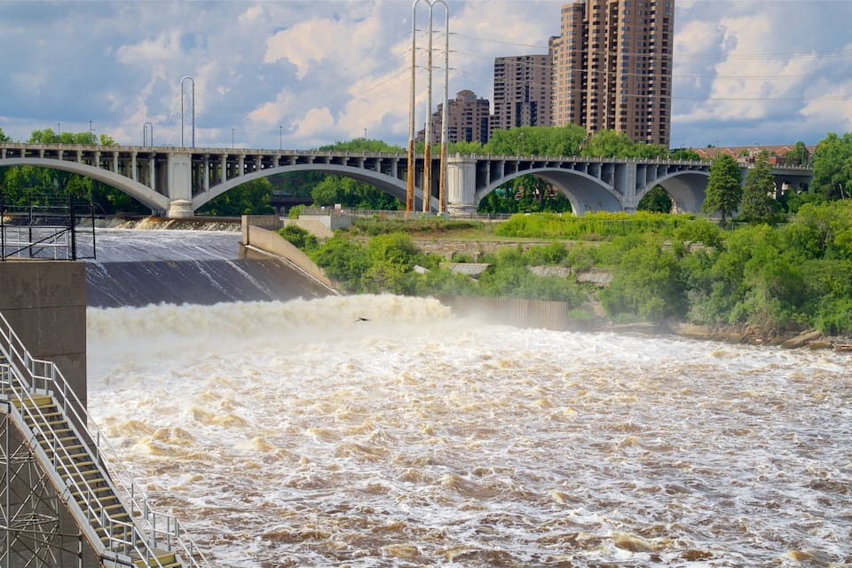 A dam with a spillway releasing water, a bridge, and high-rise buildings in the background.