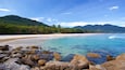 A tropical beach with clear turquoise waters, rocky shoreline, palm trees, and distant mountains.