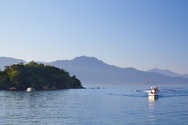 Ein ruhiges Meer mit einer kleinen, grün bewachsenen Insel und Palmen, umgeben von Bergen.