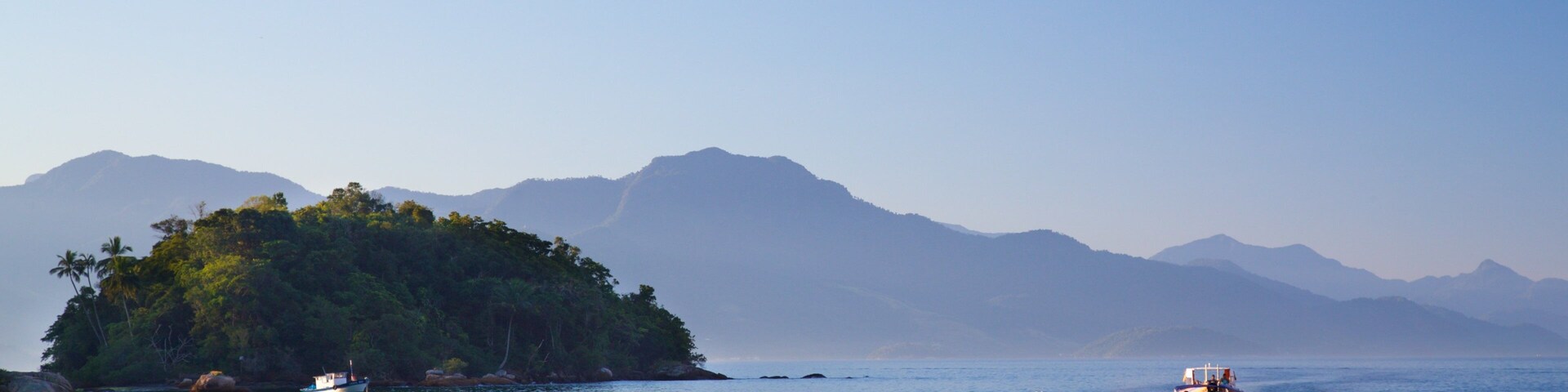 Un mar tranquilo con una pequeña isla cubierta de vegetación y palmeras, rodeada de montañas.